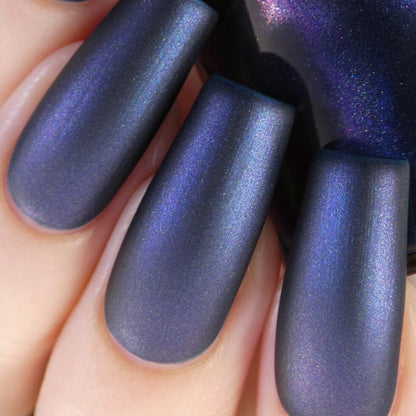 A close-up of hands with long nails painted in a deep, shimmering blue color featuring a multichrome finish shows the polish catching the light, evoking 90s nostalgia while highlighting the metallic sheen and smooth texture of PI Colors' Judge None Choose One.000 Luxury Nail Lacquer.