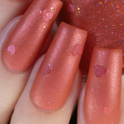 A close-up of a hand with elegantly long, glossy nails showcases the shimmery coral hue of the Strawberry Jam.228 Red Nail Polish by PI Colors. This handmade lacquer features small heart-shaped accents and is formulated as a 10-free nail polish that reflects light to create a dazzling, sparkling effect.