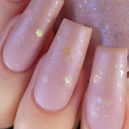 A close-up of a hand adorned with long, pink nails showcases the luxurious Special Week.000 Pink Nail Polish by PI Colors. The matte finish and hexagonal glitter pieces provide a subtle shimmer, producing a delicate and elegant style reminiscent of Uma Musume Pretty Derby.