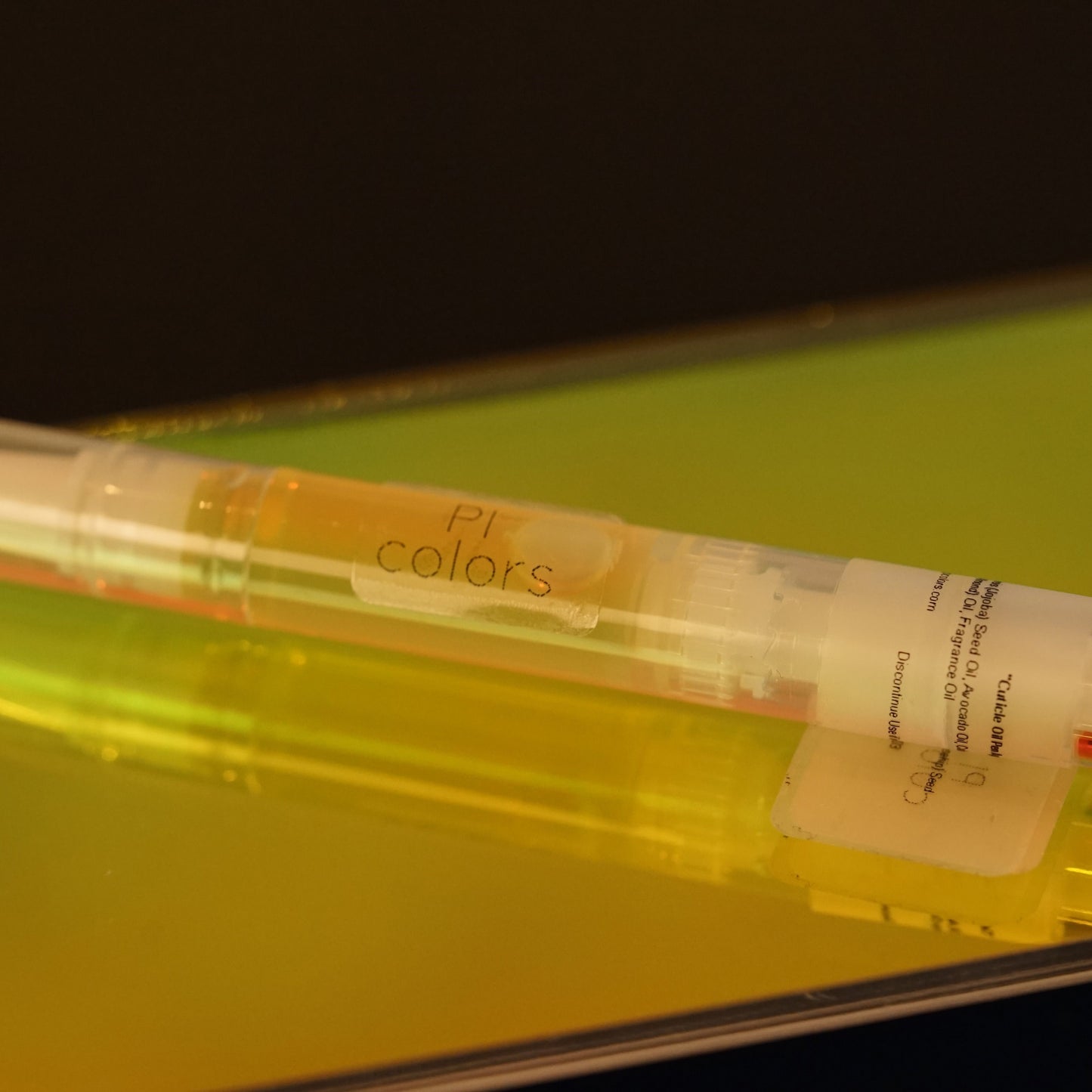 Close-up of the PI Colors "Cuticle Oil Peach Scented" product, resting on a reflective yellow and green surface. The clear container, infused with natural oils, reveals its contents inside while the softly blurred background adds an artistic touch.