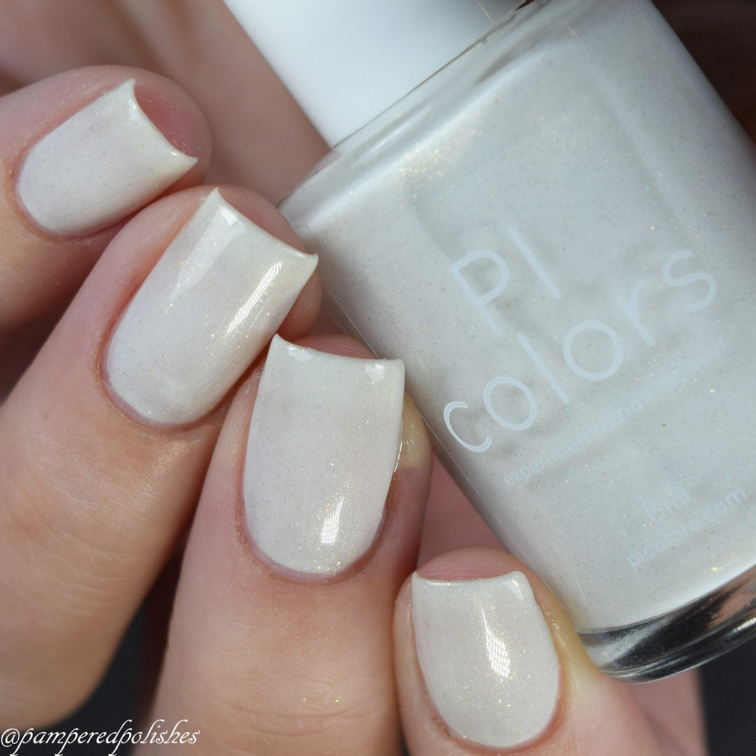 A close-up of a hand showcases nails painted in an elegant pearly white, as a bottle of "PI Colors" Aurum Dust.069 is held gracefully between the fingers. This handmade luxury nail lacquer adds a subtle shimmer and glossy finish to the nails.