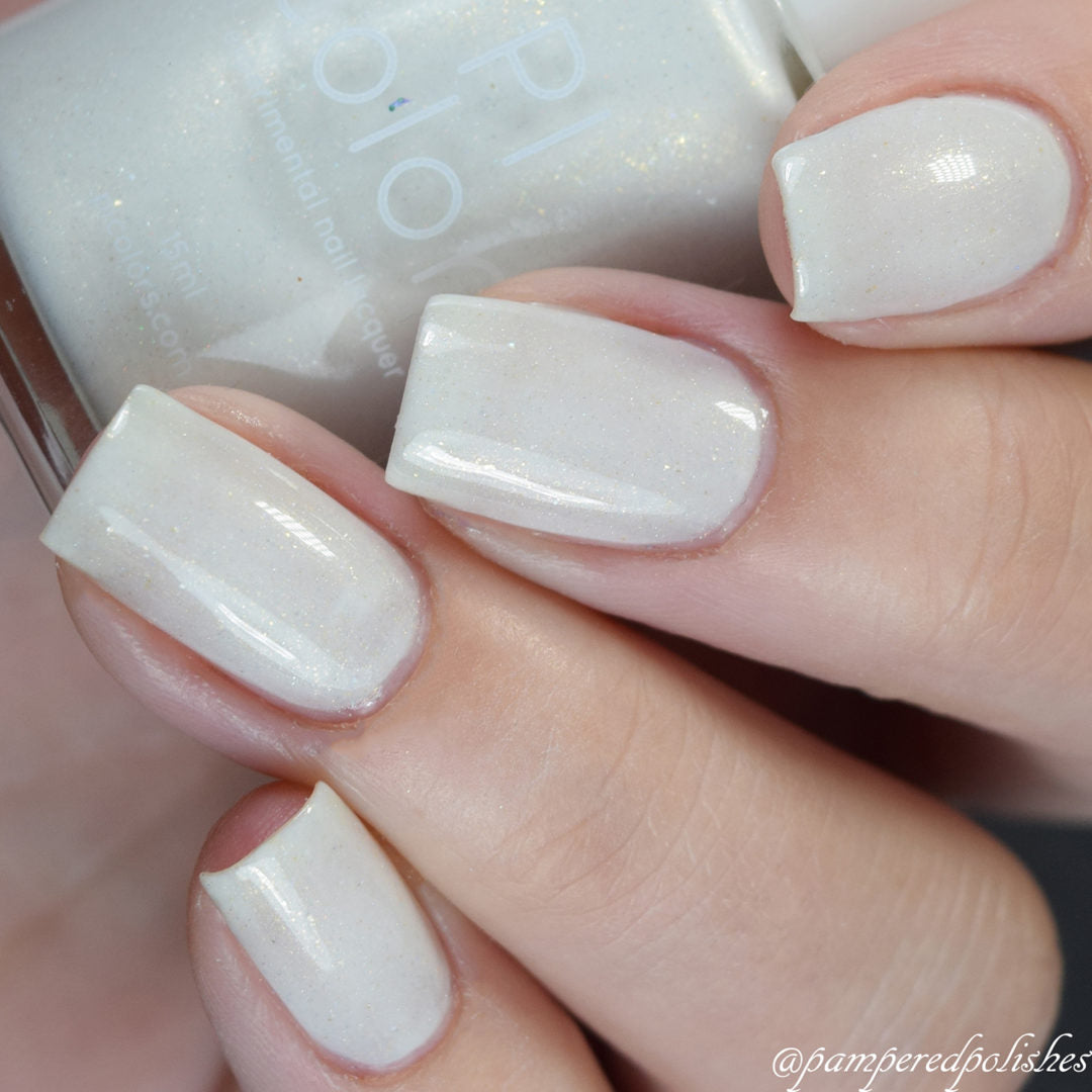 A close-up of a hand with neatly manicured nails painted in PI Colors' Aurum Dust.069 White Nail Polish, a luxurious lacquer with shimmery cream hues. The handmade polish bottle is held in the background, highlighting its color and subtle sparkle.