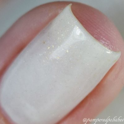 Close-up of a fingernail adorned with PI Colors' Aurum Dust.069 White Nail Polish, a luxury nail lacquer offering a glossy, pale white finish accented by subtle shimmer and glitter. The blurred background accentuates the exquisite handmade quality of this polish.