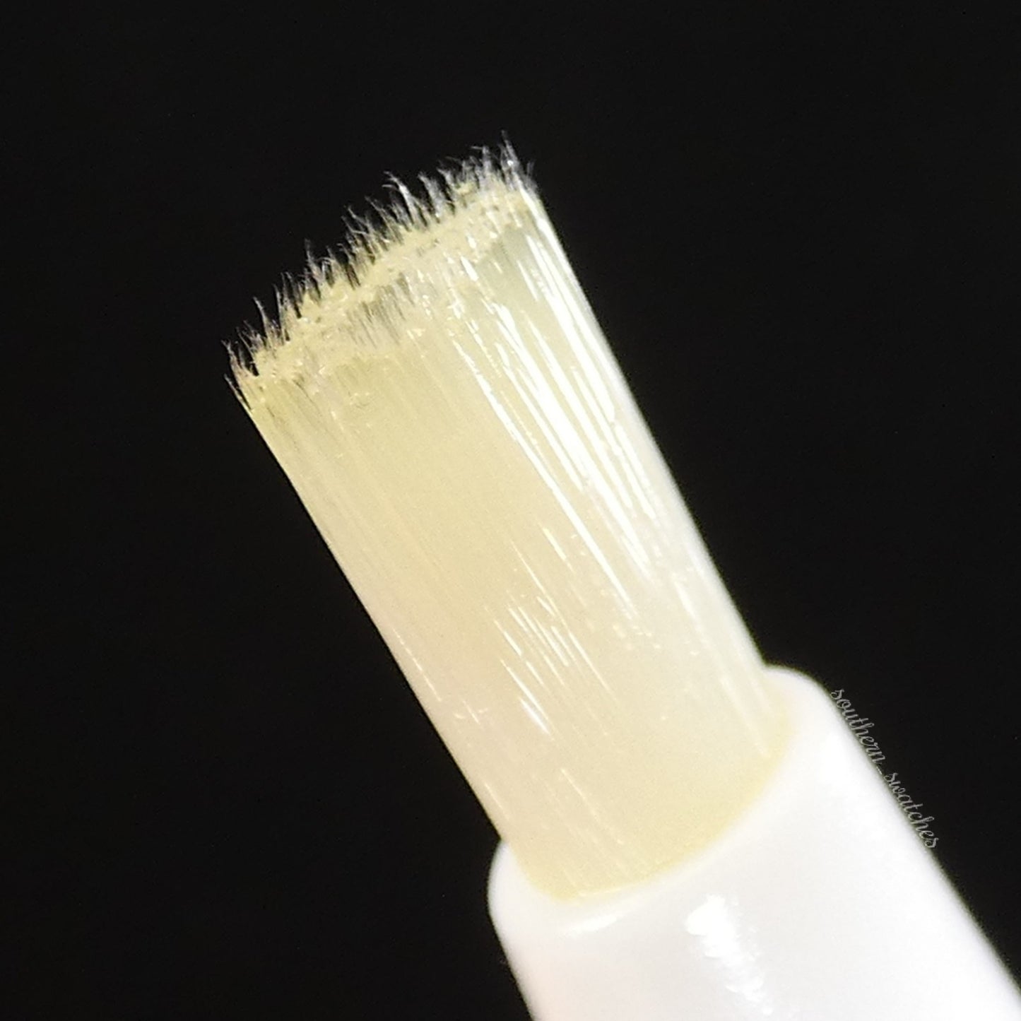 Close-up of a narrow, soft-bristled white brush against a black background, part of the PI Colors Honey Scented Cuticle Oil applicator, designed to enhance the beauty and health of your nails.