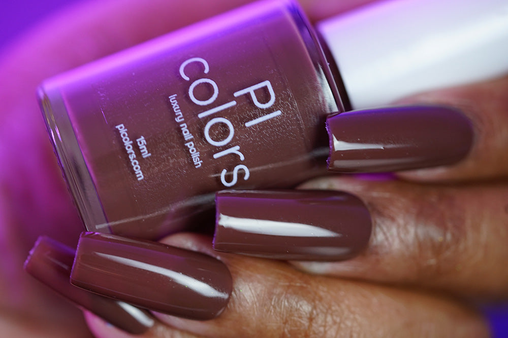 A hand with long, square-shaped nails painted in glossy Chocolate Turtle.411 by PI Colors holds a matching bottle of the luxe creme finish polish against a soft purple background.