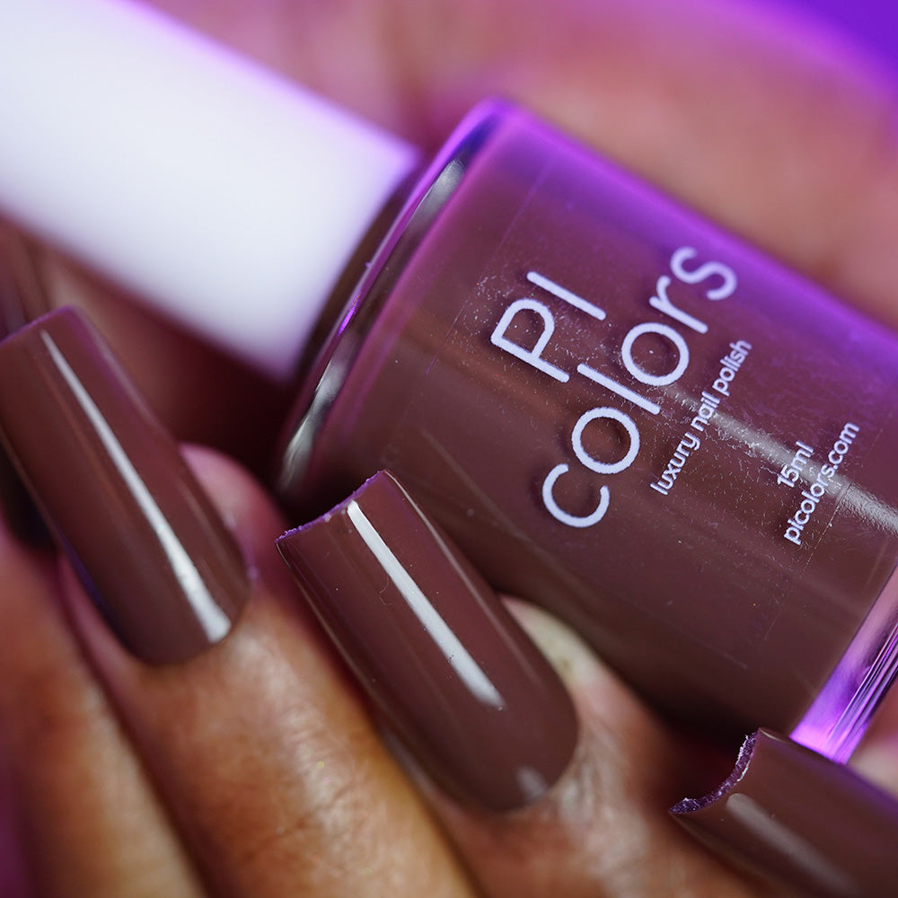 Close-up of a hand with long, glossy brown nails holding a bottle of PI Colors "Chocolate Turtle.411" handmade nail polish. The polish features a smooth, shiny creme finish that matches the nails perfectly.