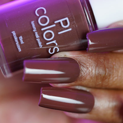 A close-up of a hand with long, square-shaped nails painted in Chocolate Turtle.411, a glossy deep mauve crème by PI Colors, holding its matching handmade nail polish bottle.