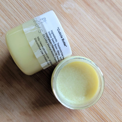 A small open jar of PI Colors Cuticle Butter sits on a wooden surface. The creamy, pale yellow texture gives off a hint of chocolate scent. Its label highlights organic ingredients like shea and aloe butters. The lid, resting on its side, displays additional text and brand information.