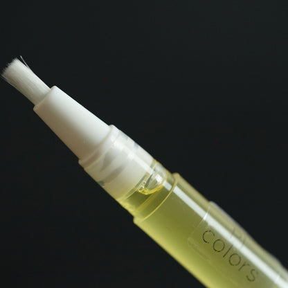 A close-up of the PI Colors Cuticle Oil Peach Scented pen showcases its brush tip. The pen, filled with a yellow liquid enriched with jojoba and tamanu oils, features a clear plastic body that gleams against the dark background, making it stand out vividly.