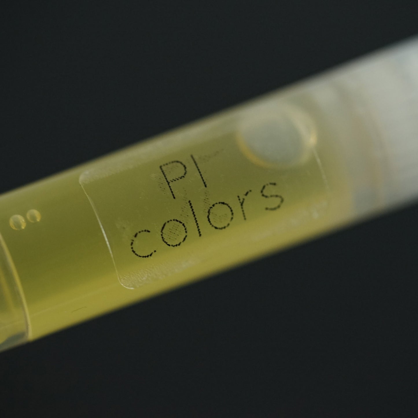 A close-up of a test tube filled with yellow liquid, labeled "PI Colors," suggests the presence of natural oils such as jojoba and tamanu. The horizontally positioned tube against a dark background encapsulates the essence of PI Colors' Peach Scented Cuticle Oil.