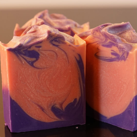 The Autumn Moon Artisan Soap Bars by PI Colors feature intricate purple and orange swirls with spiced orange scent, crafted from natural ingredients. The design boasts artistic flair atop a plain backdrop.