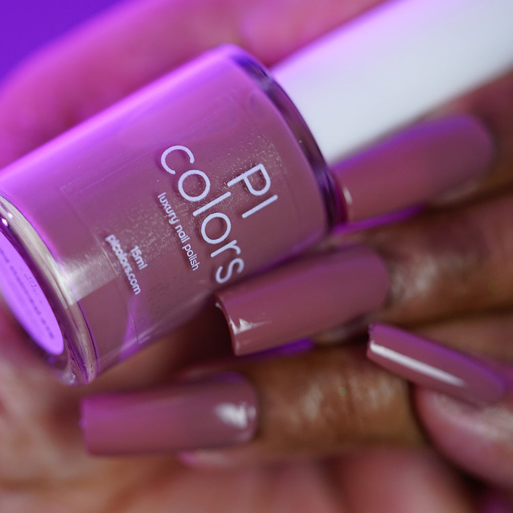 A hand with long, glossy nails painted in PI Colors’ Iced Oatmeal.478 holds the matching bottle under purple lighting, highlighting its luxurious creme finish.