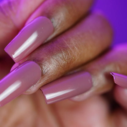 A close-up of a hand with long, square acrylic nails painted in PI Colors' Iced Oatmeal.478, a glossy mauve creme. The pinky nail is chipped at the tip; the background is blurred purple.