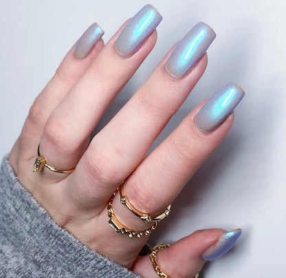 A close-up shot highlights a hand adorned with long, luxurious nails painted in Cloud Rainbow.303 Blue by PI Colors, featuring mesmerizing color-changing effects. The opulent look is further enhanced by multiple gold rings, while a gray garment sleeve subtly complements this splendid display.