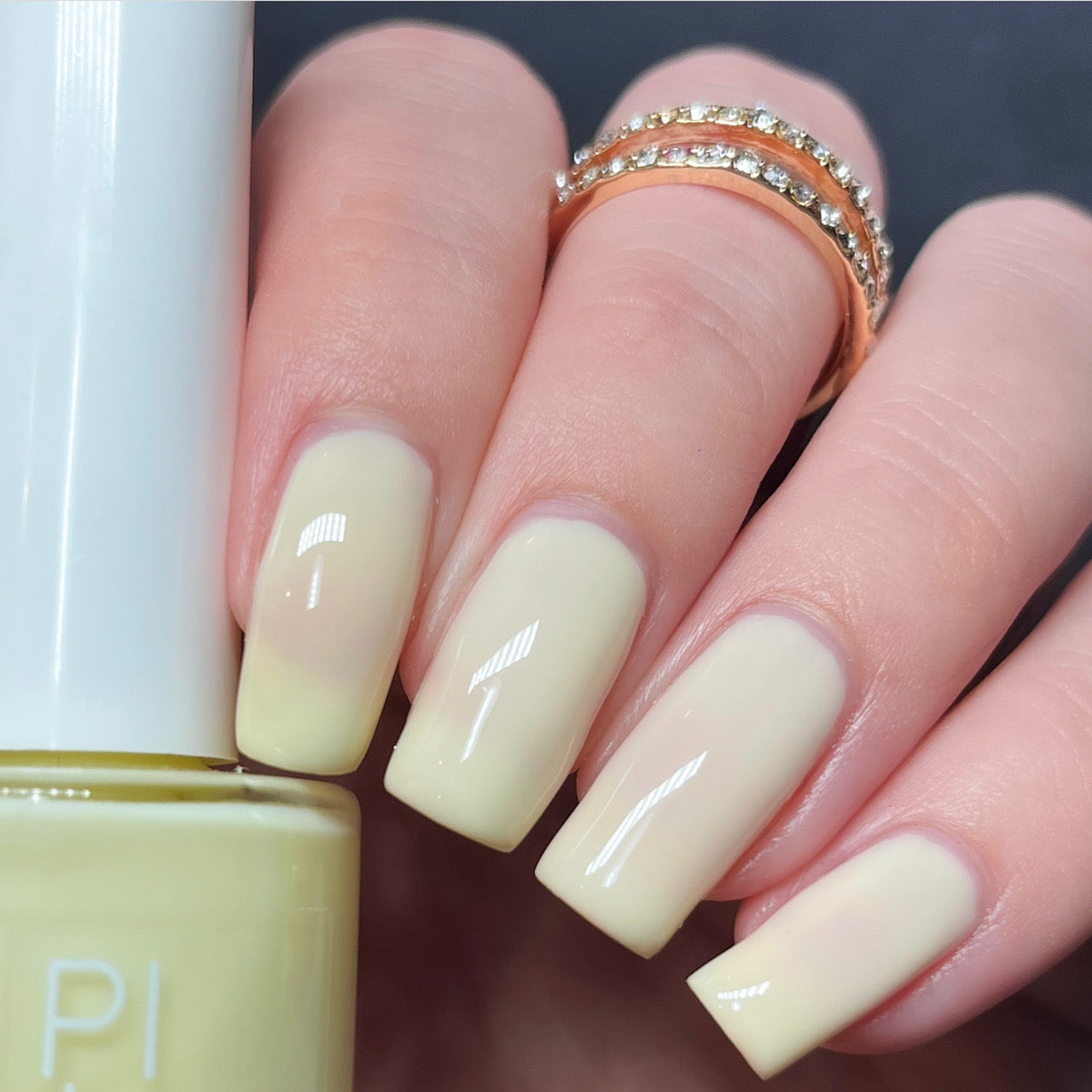 A hand with neatly manicured nails painted in the glossy, pastel shade of Chiffon.400 Yellow Nail Polish from PI Colors exudes elegance. The look is further accentuated by two stacked gold rings adorned with shimmering stones, while a bottle of this creme finish nail polish is partially visible in the foreground.