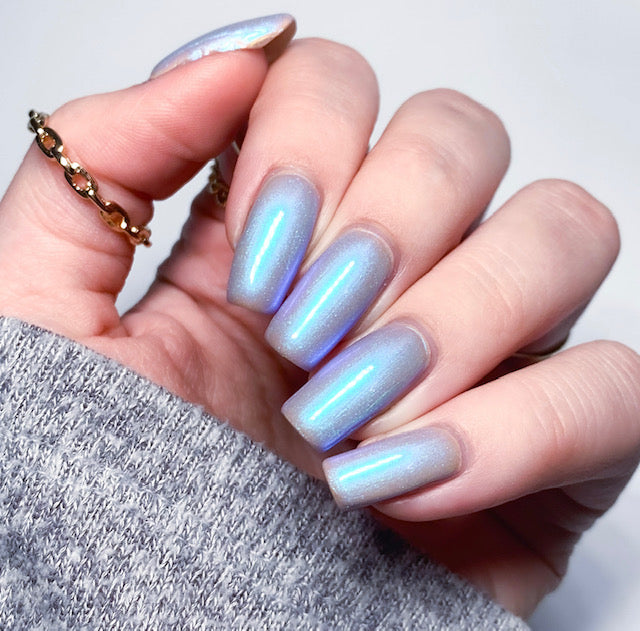 A hand with long, shiny nails showcases the opulence of PI Colors' Cloud Rainbow.303 Blue Nail Polish, revealing holographic shades of blue and purple. The person wears a gray sleeve and a gold ring adorned with small stones, set against a soft, neutral backdrop.