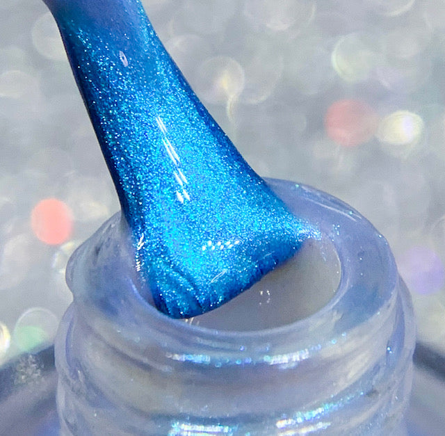 An up-close view of a brush showcases the vibrant and luxurious Cloud Rainbow.303 Blue Nail Polish by PI Colors in an electric silver blue shade. The shimmering texture stands out against the blurred, glittery background, emphasizing its vivid color-changing effects and glossy finish.