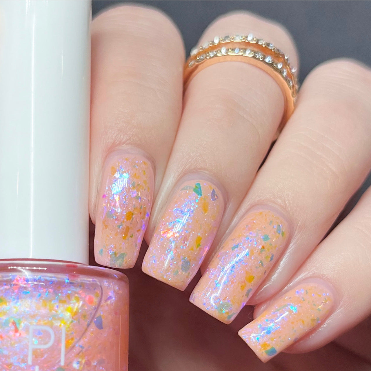 A close-up of a hand with nails painted in PI Colors' Pink Sapphire.404 Pink Nail Polish Topper, showcasing a color-shifting flakie effect. The fingers are adorned with two gold rings featuring small diamonds, and a matching nail polish bottle sits elegantly alongside the display.