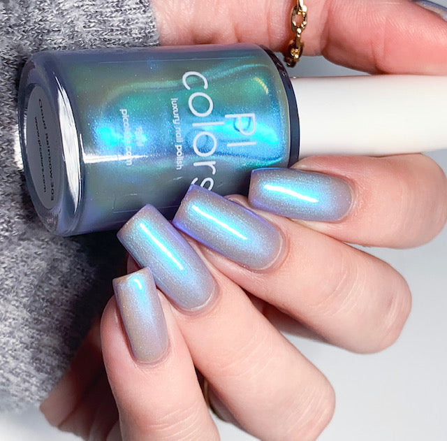 A hand with long, glossy nails in a colorshifting blue shade holds a bottle of PI Colors' Cloud Rainbow.303 Blue Nail Polish, which shimmers with a reflective quality. The person is wearing a grey long sleeve and a thin gold ring on one finger, adding an elegant touch to the chic ensemble.