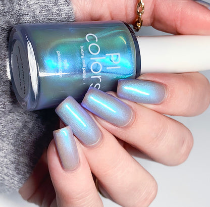 A hand with long, glossy nails in a colorshifting blue shade holds a bottle of PI Colors' Cloud Rainbow.303 Blue Nail Polish, which shimmers with a reflective quality. The person is wearing a grey long sleeve and a thin gold ring on one finger, adding an elegant touch to the chic ensemble.