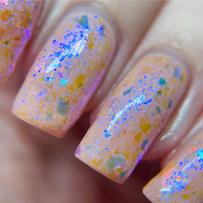 A close-up shot showcases fingernails embellished with PI Colors’ Pink Sapphire.404 Pink Nail Polish Topper, an iridescent, multicolored confetti-style lacquer. This color-shifting flakie topper reflects blue, orange, and green hues over a peach-colored base for a dazzling effect.