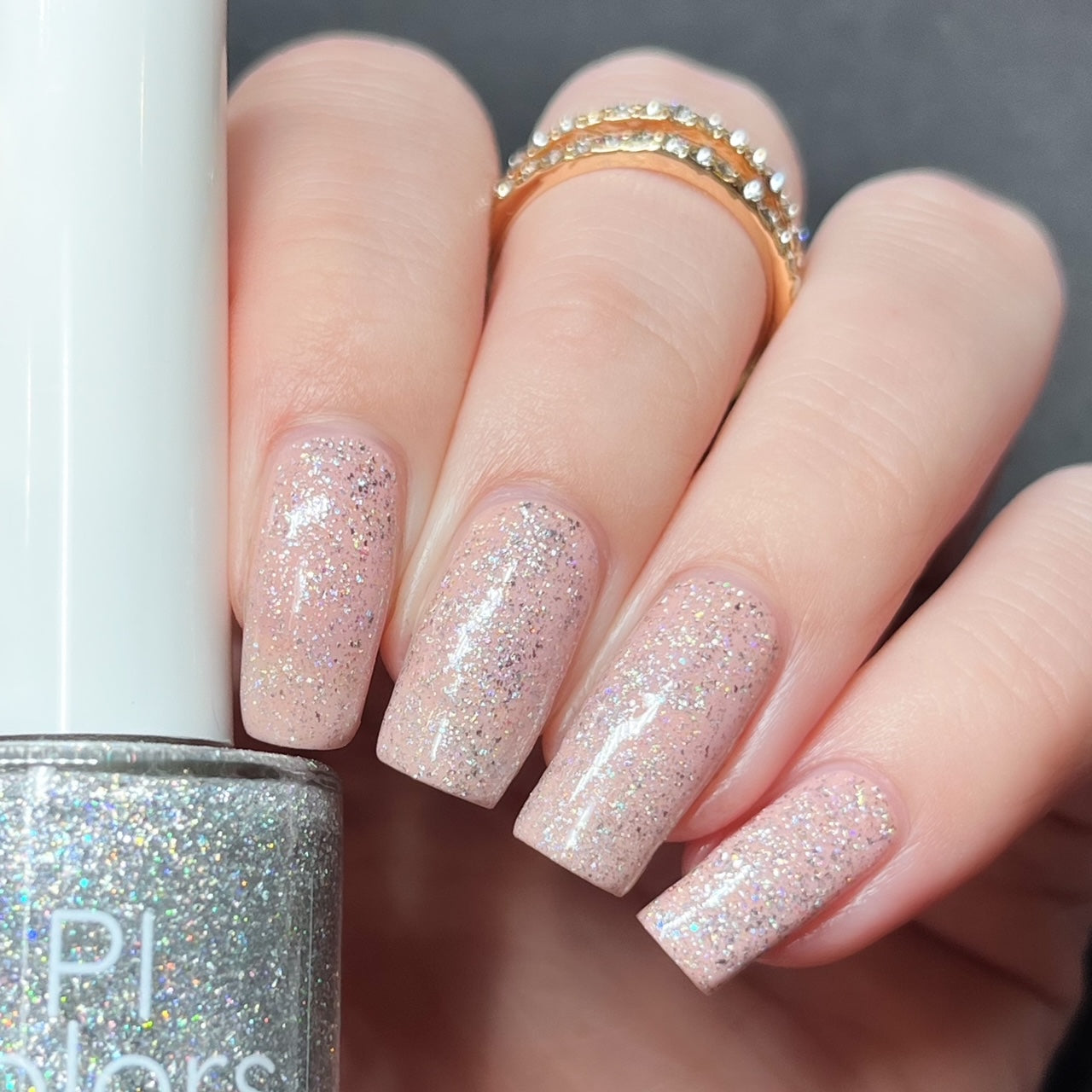 A hand with long, square-shaped nails flaunts a layer of PI Colors' "Diamond Blast.401," a luxury nail lacquer that adds a glittery, pale pink finish enhanced by silver holographic glitter. The partially visible bottle, capped in white, showcases this exquisite polish topper. A gold ring adorned with small beads completes the elegant look against the solid gray background.