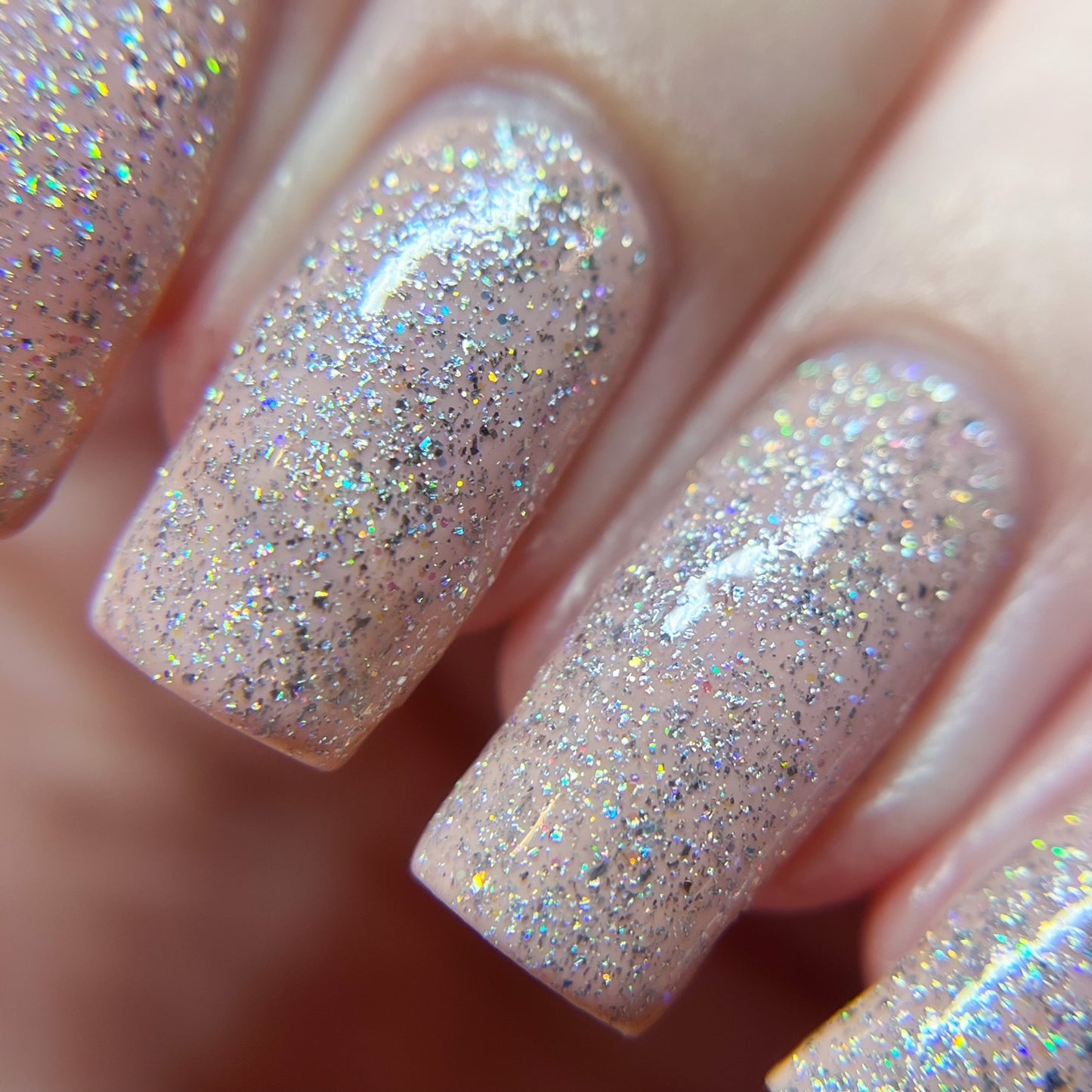 A close-up showcases fingers adorned with nails painted in a luxury nude polish, topped with PI Colors' Diamond Blast.401 Silver Holographic Glitter Nail Polish Topper. The fine glitter catches the light, creating a shimmering effect that highlights the smooth texture and clean edges of the nails.