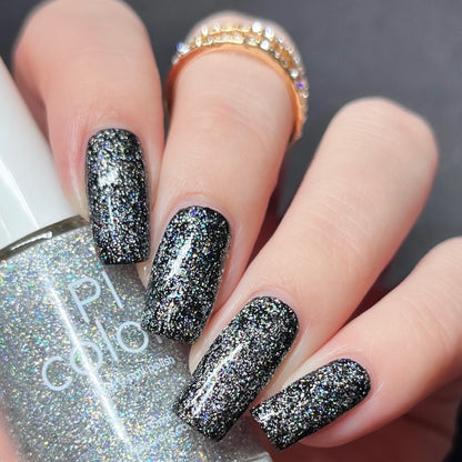 Close-up of a hand with long nails painted in luxury nail lacquers featuring glossy black and the Diamond Blast.401 Silver Holographic Glitter Nail Polish Topper from PI Colors. The hand holds a bottle of similar 10-free nail polish. A gold ring with small gems adorns the middle finger, completing the elegant look.