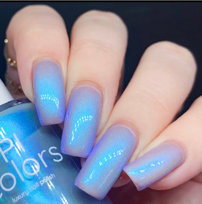 A close-up of a hand with long, square-shaped nails showcases the luxurious Cloud Rainbow.303 Blue Nail Polish by PI Colors in an electric silver blue shade, featuring a subtle gradient effect. The partially visible bottle is elegantly labeled "PI Colors," suggesting its mesmerizing colorshifting allure.