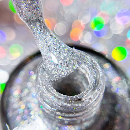 A close-up of a PI Colors nail polish brush dripping Diamond Blast.401 Silver Holographic Glitter Nail Polish Topper into an open bottle, set against a softly blurred background with colorful, shimmering bokeh lights that create a sparkling and festive atmosphere, ideal for showcasing luxury nail lacquers.