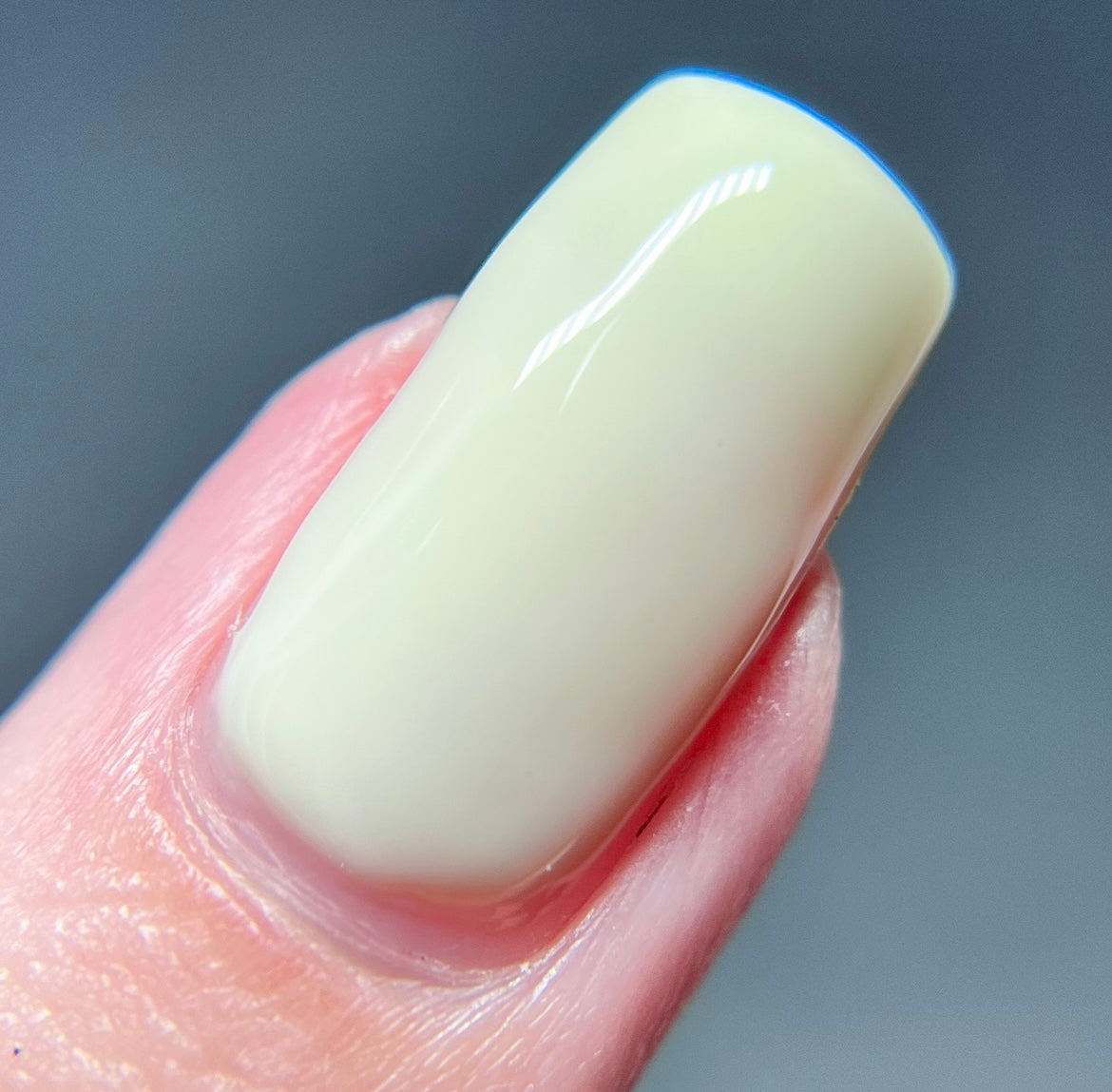 Close-up of a fingernail painted in Chiffon.400, a glossy, pastel yellow shade from PI Colors. The creme finish of this Yellow Nail Polish leaves the surrounding skin looking smooth, set against a blurred gray background.