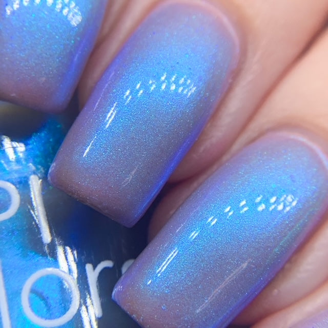 Close-up of fingers adorned with the luxurious Cloud Rainbow.303 Blue Nail Polish by PI Colors, showcasing a shimmering effect. The iridescent hue beautifully reflects light, revealing subtle color-changing effects. A bottle of this handmade nail polish is partially visible in the background.