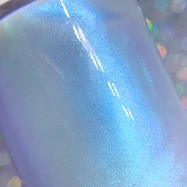 A close-up showcases the smooth, luxurious polish of PI Colors' Cloud Rainbow.303 Blue Nail Polish, its iridescent finish shifting between shades of blue and purple. The background features a soft-focus effect with multicolored light spots that create a dreamy and ethereal atmosphere, enhanced by color-changing effects.