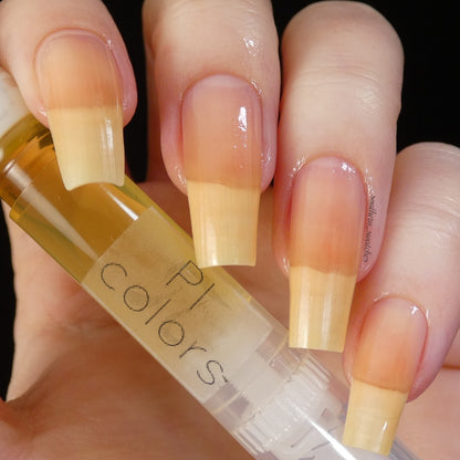 A close-up image of a hand with long, beige-gradient nails holding a small clear bottle labeled "PI Colors," showcasing the nourishing blend of jojoba oil. The bottle contains a light yellow liquid, ideal for cuticle care, and is named Honey Scented Cuticle Oil. The background is black.