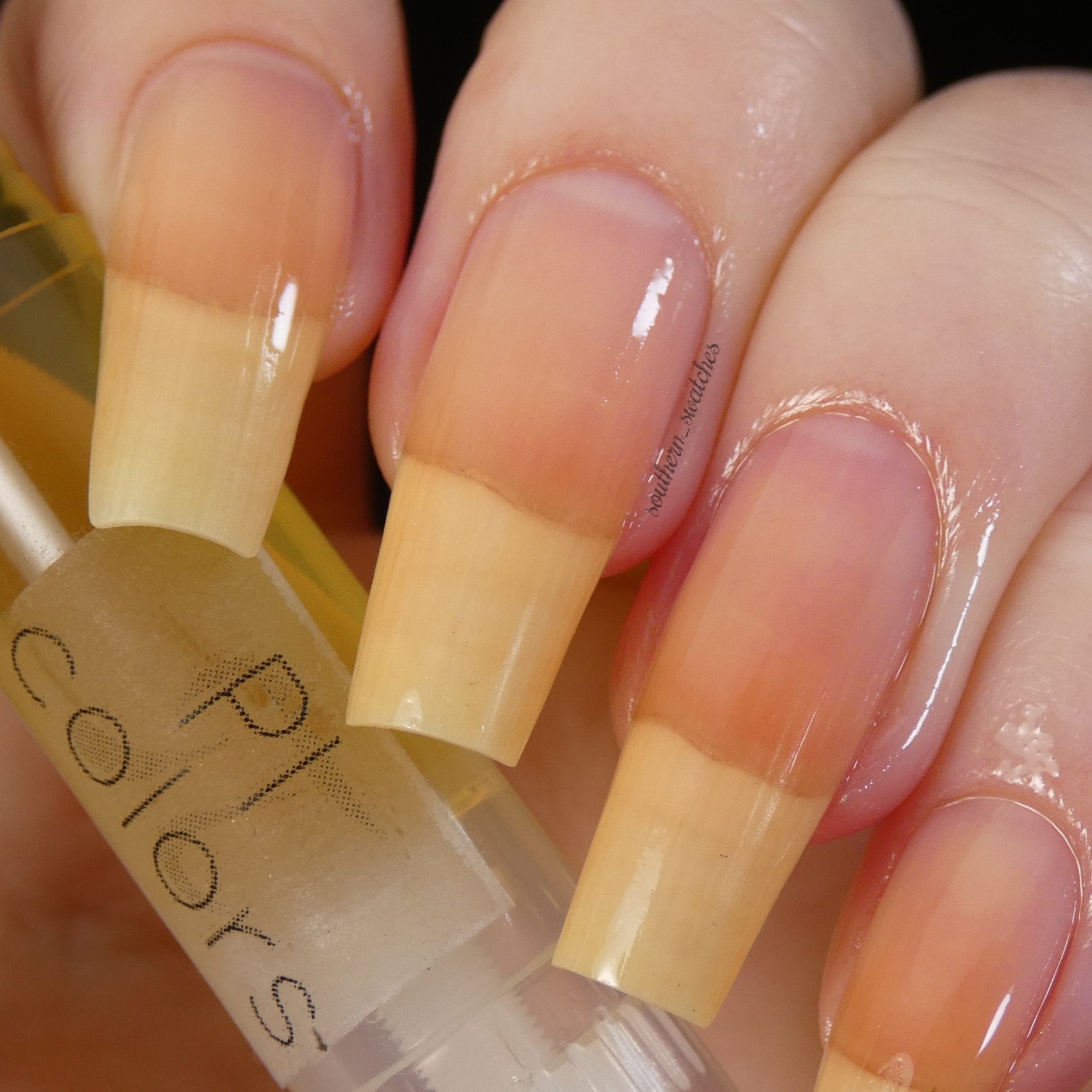 A close-up image showcases a hand with long, natural nails featuring a gradient from light pink at the base to yellow at the tip. The hand is holding a transparent bottle of PI Colors' Honey Scented Cuticle Oil, with part of the label visible. The nails look glossy and well-groomed, highlighting the nourishing effects of jojoba oil.