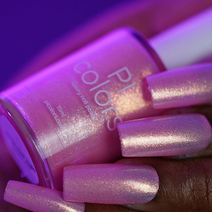 A hand with long, square nails painted in PI Colors’ Lemon Pink Sugar Cookie.422—a shimmery, iridescent light pink—holds the matching bottle under purple lighting, highlighting the luxurious appeal of this nail lacquer.