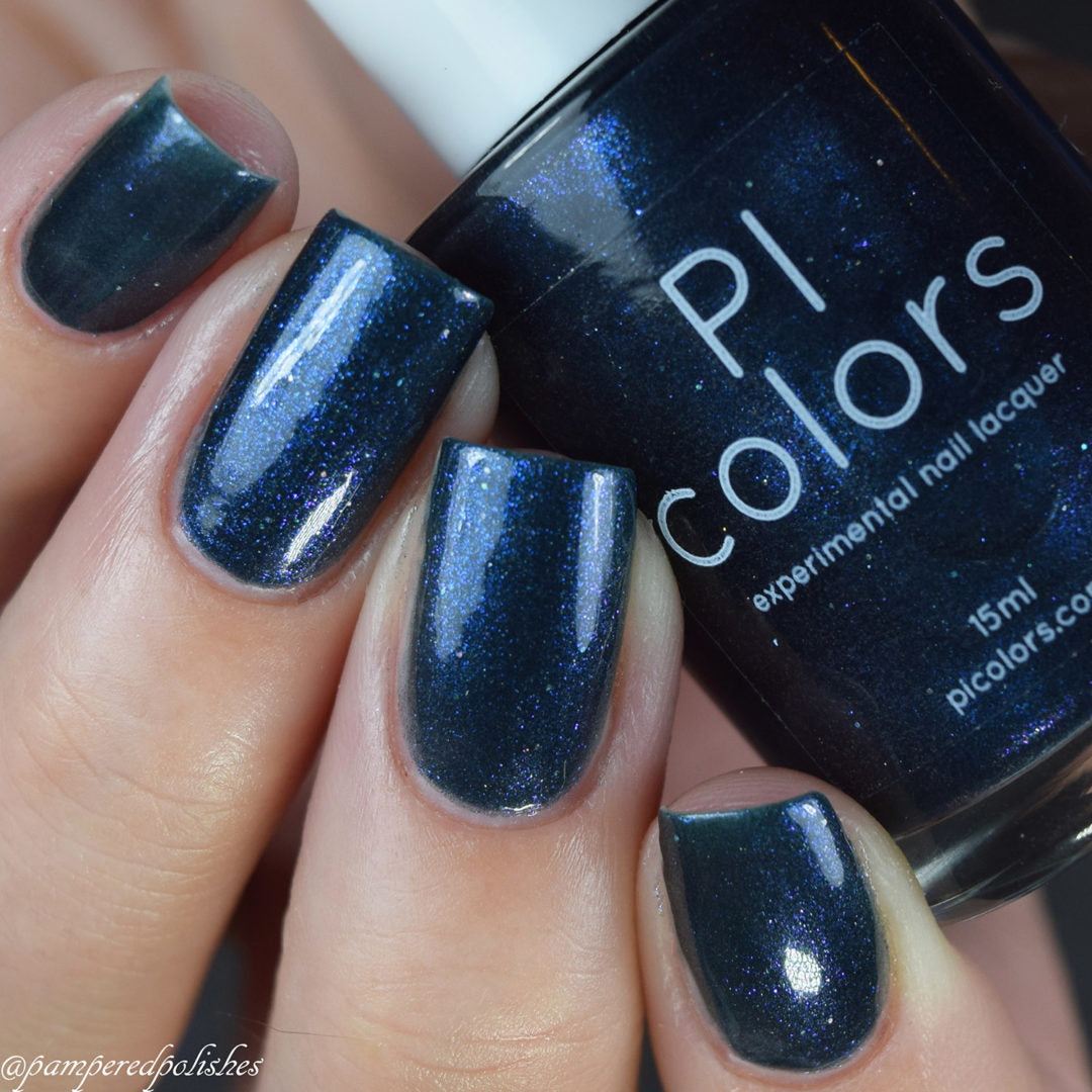 Close-up of a hand with nails painted in "Midnight Bloom.022," a shimmering blue-black nail polish by PI Colors. The bottle, partially visible, reveals the brand’s signature style. This blue-tinted polish offers a glossy finish adorned with delicate specks of glitter, capturing attention with its radiant sparkle.