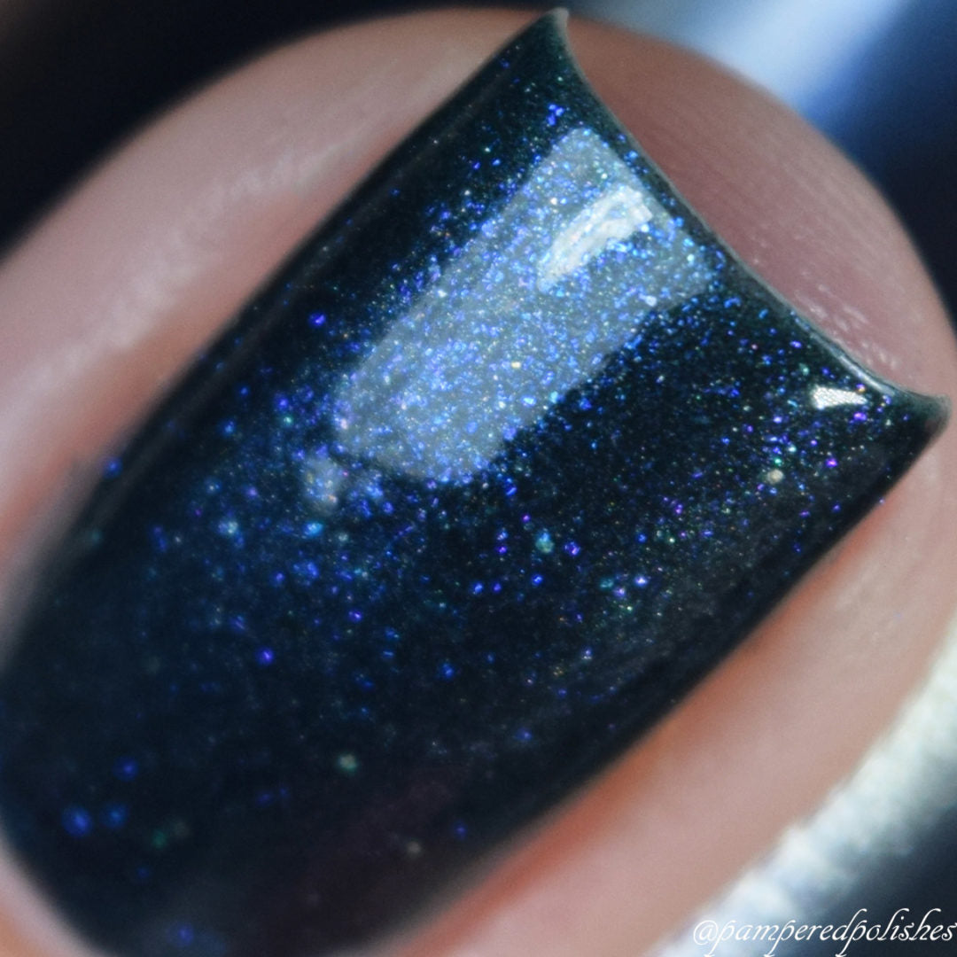 Close-up of a fingernail painted with PI Colors' Midnight Bloom.022 Blue Black Nail Polish, showcasing an enchanting blue shimmer and glitter finish. The polish reflects light, producing a sparkling effect.