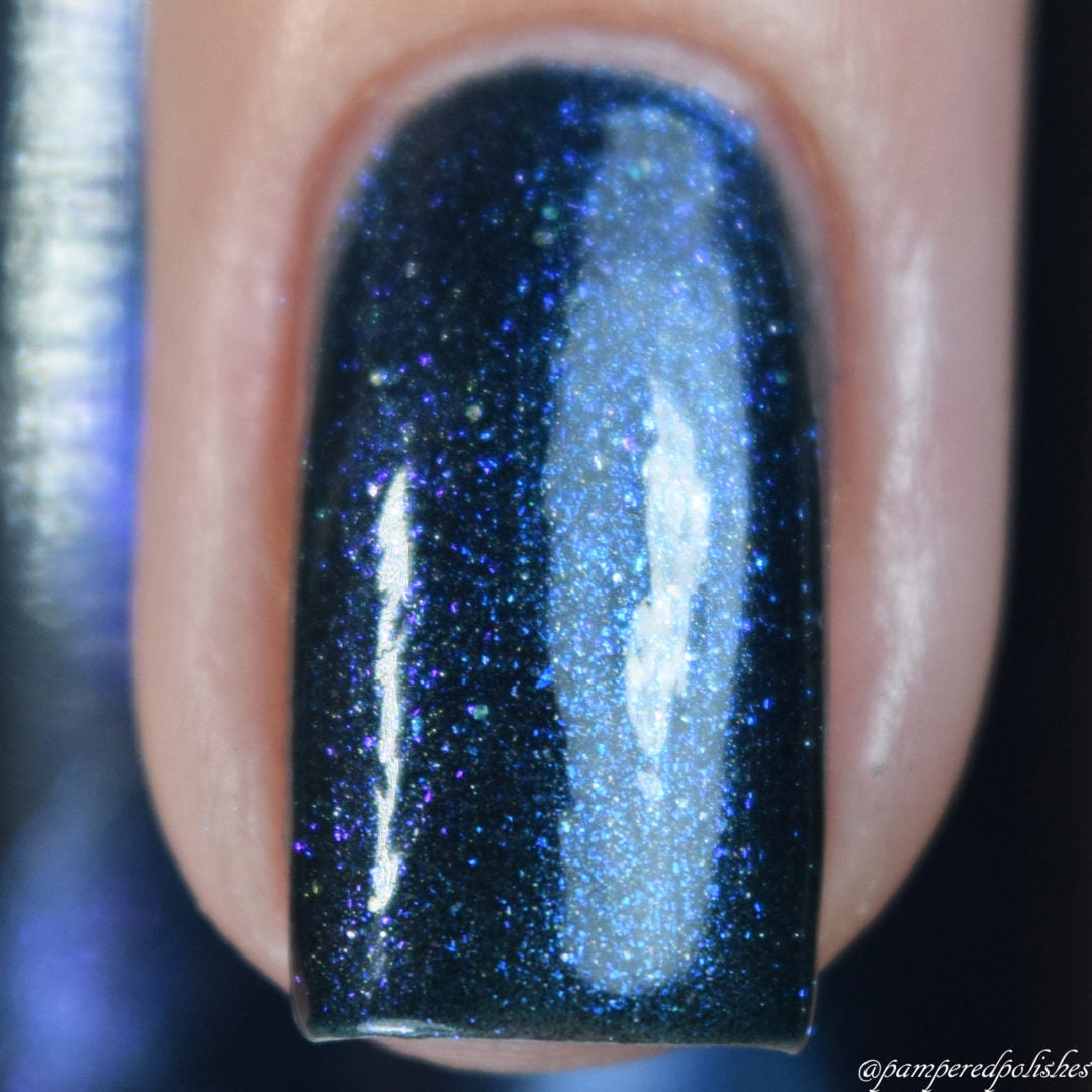 A close-up of a fingernail adorned with PI Colors’ Midnight Bloom.022 Blue Black Nail Polish showcases a shimmering dark blue and purple holographic effect. The glossy polish, speckled with glitter, reflects light beautifully for a radiant look, while subtle hints of black add depth to this mesmerizing display.