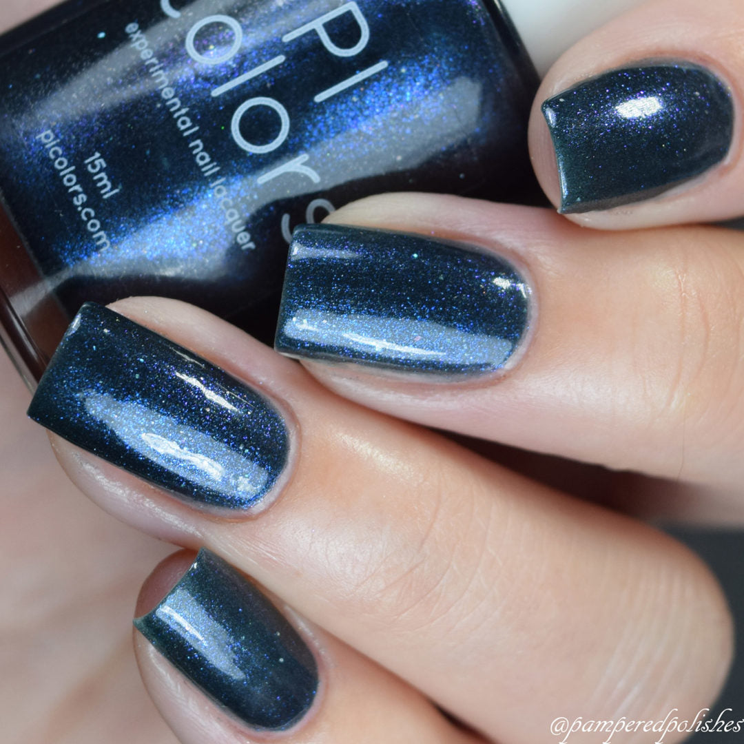 A hand showcases nails adorned with the deep blue sheen of Midnight Bloom.022 Blue Black Nail Polish, glinting brilliantly under the light. The bottle, branded as "PI Colors," is partially visible in the background, revealing a glossy and iridescent finish with captivating hints of glitter.