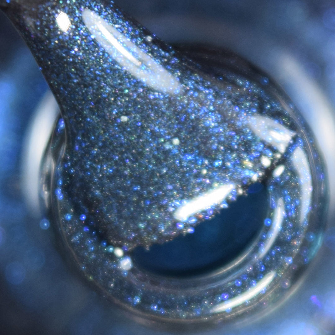 Close-up of the Midnight Bloom.022 Blue Black Nail Polish brush from PI Colors, shimmering with glitter and hints of black polish, poised above the open neck of the bottle. The image captures the dynamic texture and reflective sparkle of the blue shimmer under light.