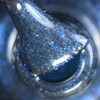 Close-up of the Midnight Bloom.022 Blue Black Nail Polish brush from PI Colors, shimmering with glitter and hints of black polish, poised above the open neck of the bottle. The image captures the dynamic texture and reflective sparkle of the blue shimmer under light.