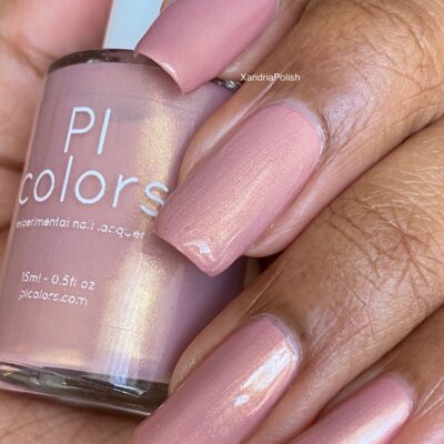 A close-up shows a hand adorned with long, manicured nails, painted in a shimmering nude pink. The polished nails grasp a coordinating bottle of PI Colors' Milk Berry.073 Pink Nail Polish, which boasts a luxurious, glossy finish under the light.