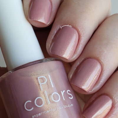 A close-up shot highlights a hand with short nails adorned in a shimmery, muted pink shade, capturing the luxurious appeal of PI Colors' Milk Berry.073 Nail Polish. The hand holds a bottle of the handmade polish labeled "PI Colors," perfectly matching the elegant hue on the nails and exuding sophistication and style.