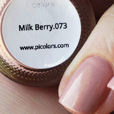 A close-up of a hand holding a jar of PI Colors' luxury Milk Berry.073 Pink Nail Polish reveals its shimmering pink shade applied on one fingernail. The label displays its name along with the website address, www.picolors.com.