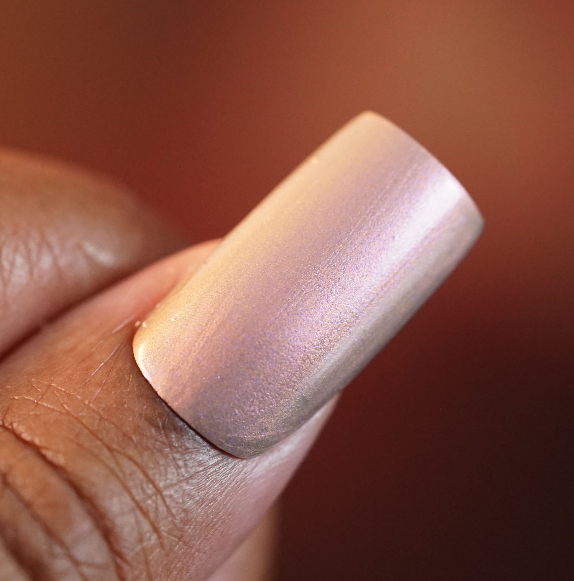 Close-up of a person's fingernail painted with Mulberry Love.205 by PI Colors, a luxury nail lacquer that shifts between soft pink and gold tones. A softly blurred background in warm hues highlights the elegance of this handmade polish.