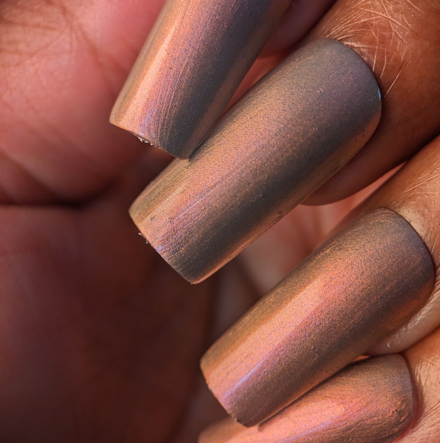 A hand with long nails is adorned in PI Colors' Mulberry Love.205, a luxurious iridescent polish shifting between pink, copper, and purple hues. The smooth, glossy finish reflects light beautifully, showcasing stunning color variations.