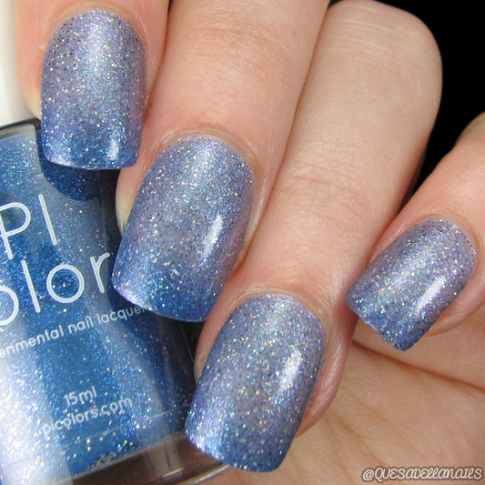 Close-up of a hand with four fingernails painted in PI Colors' Cockatoo.036 Blue Nail Polish, a 10-free formula. The partially visible bottle shows off the same sparkly blue shade. The glittery finish reflects light beautifully, creating a shimmering effect that combines luxury with safe nail care.