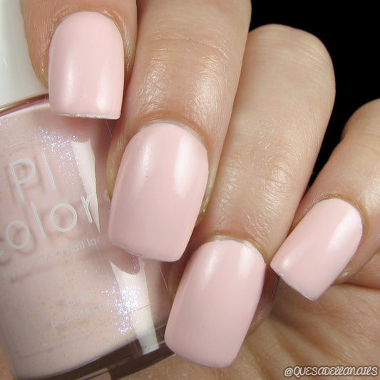 A close-up of a hand with neatly manicured nails adorned in Pink Lux.005, a soft pink shimmer crème nail polish. The bottle of the nail polish is partially visible, labeled with "PI Colors.