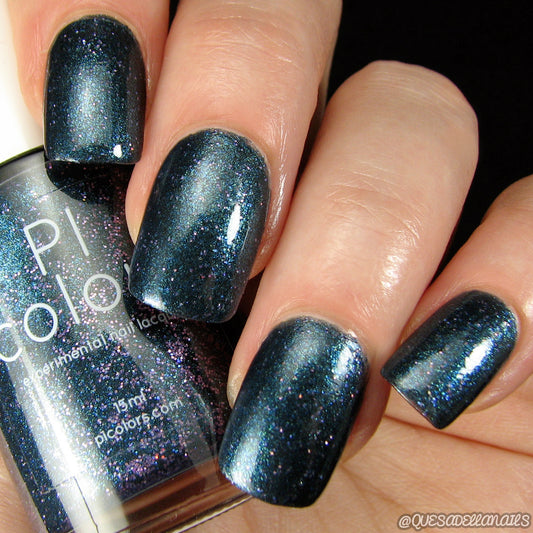 A close-up of fingernails painted with Watermelon Rind.056 Black Nail Polish by PI Colors showcases a dark, shimmery blue hue with subtle holographic glitters. The nails are neatly manicured, and the polish has a glossy finish. A 10-free bottle of the same nail polish is visible in the background.